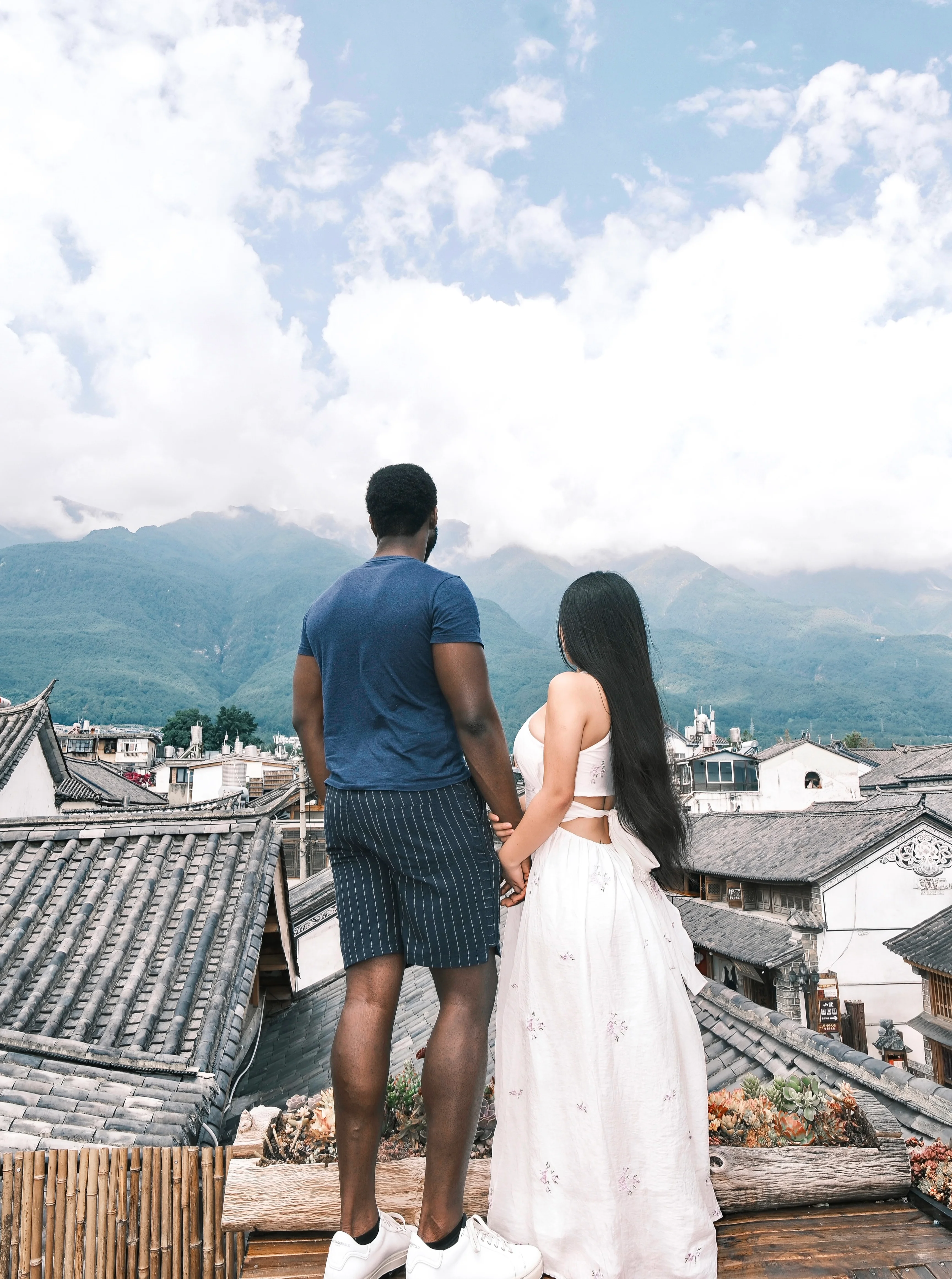 Theophilus and his wife seen from behind on a rooftop, holding hands and looking out across tiled roofs toward cloud-covered mountains